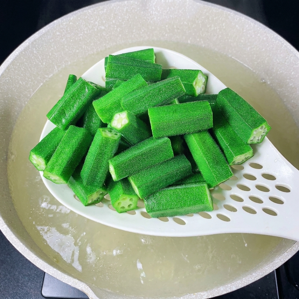 Ein weißer Schaumlöffel hebt blanchierte Okra aus dem Wasser.