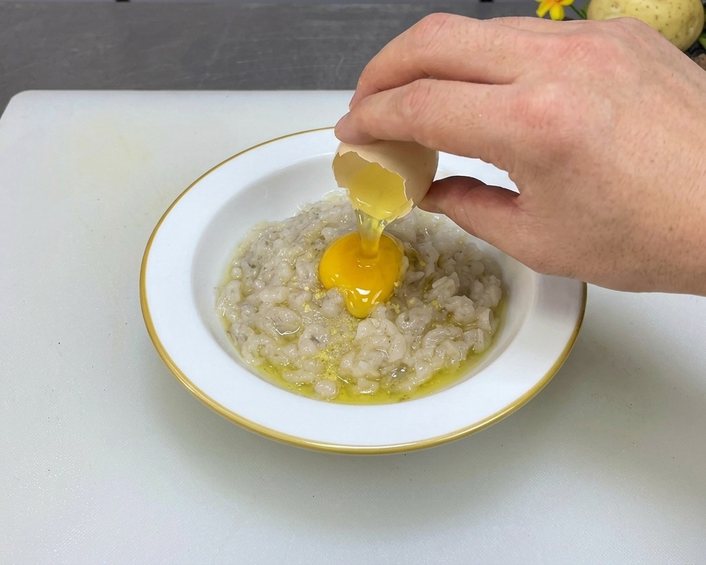 Eine Hand schlägt ein frisches Ei über einer Schüssel mit gewürzter grauer Garnelenpaste auf.