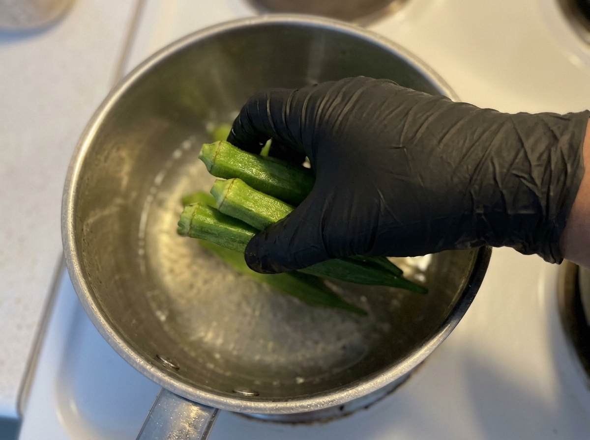 Eine Hand gibt frische grüne Okraschoten in einen Topf mit kochendem Wasser.