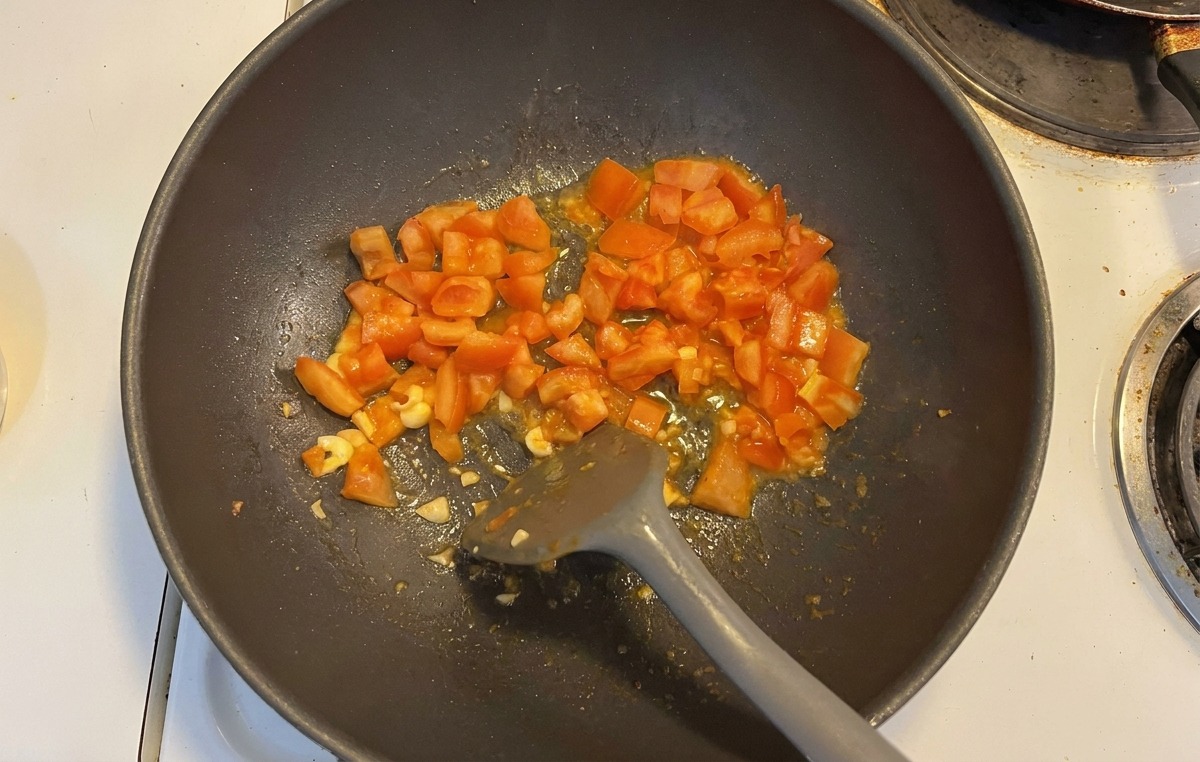 Tomatenwürfel und Knoblauchscheiben werden in einem dunklen Wok angebraten.