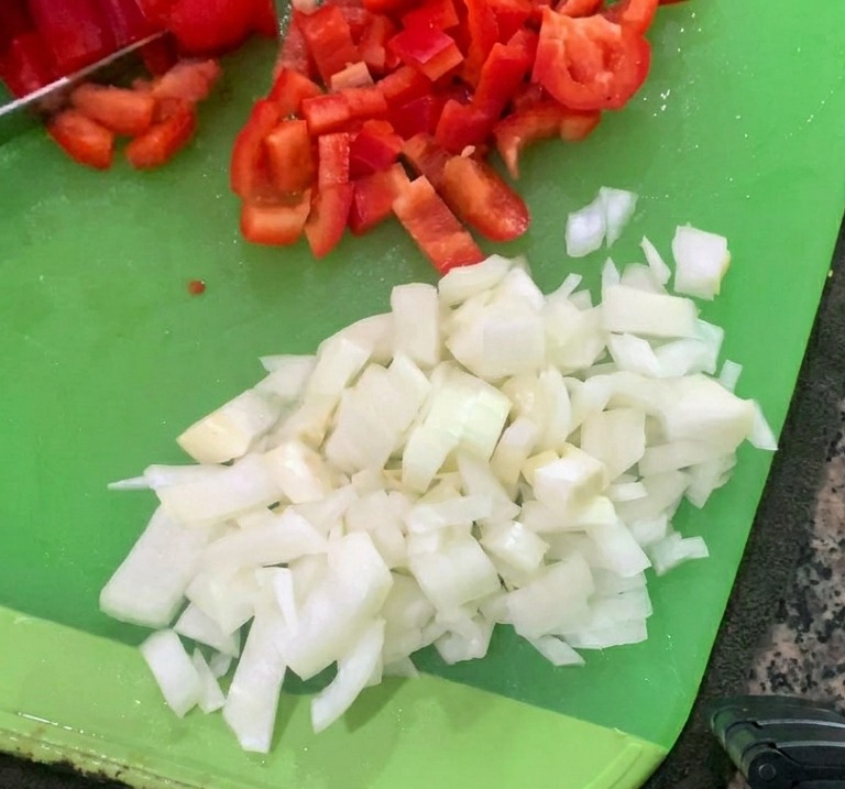 Gewürfelte Zwiebeln und gehackte rote Paprika liegen auf einem Schneidebrett.