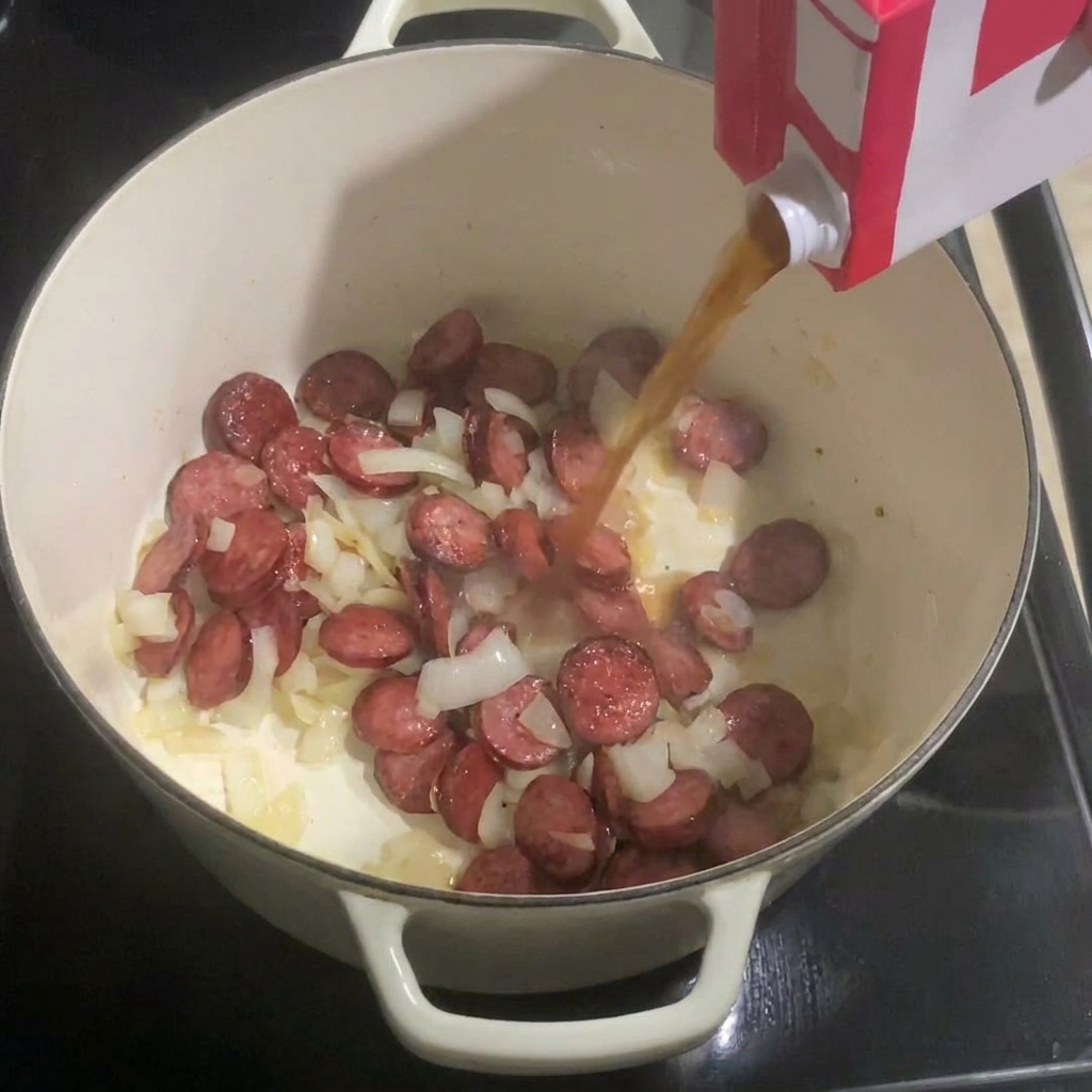 Hühnerbrühe wird aus einer Packung in einen Topf mit Wurst und Zwiebeln gegossen.