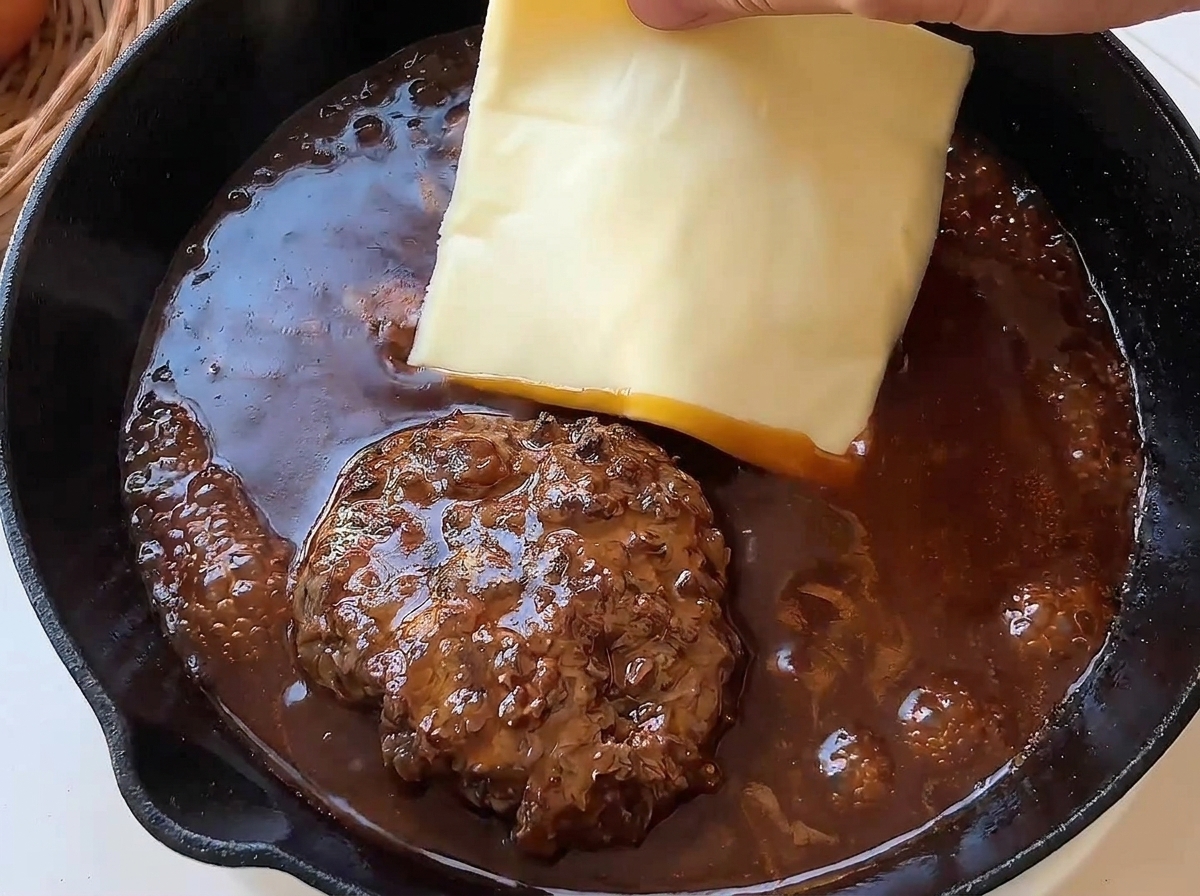 Eine Hand legt eine Scheibe Käse auf ein heißes Patty in brauner Sauce.