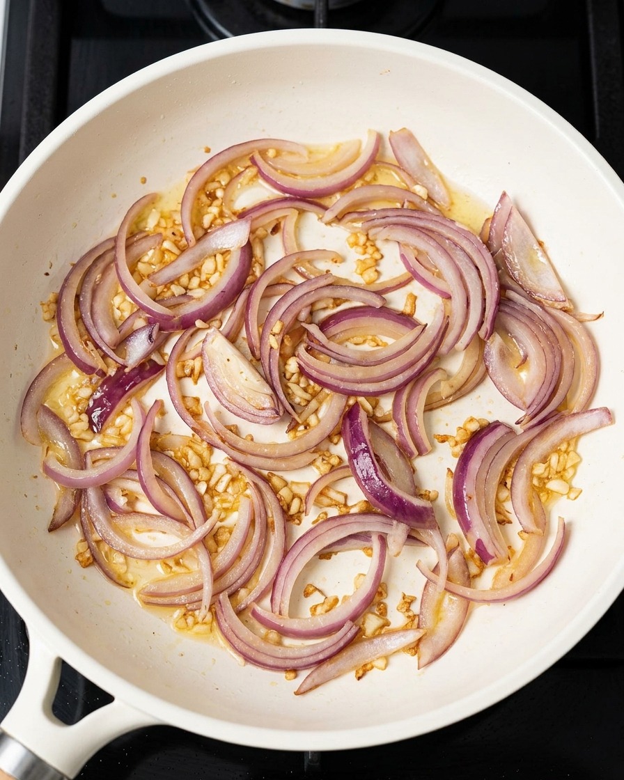 Rote Zwiebelstreifen und gehackter Knoblauch dünsten in einer Pfanne mit restlichem Öl.