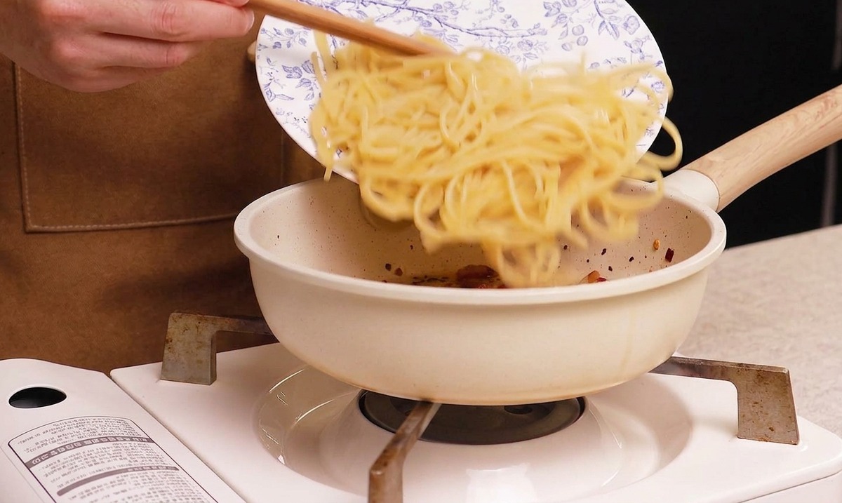 Gekochte Spaghetti werden von einem Teller in die Pfanne zum knusprigen Guanciale gegeben.
