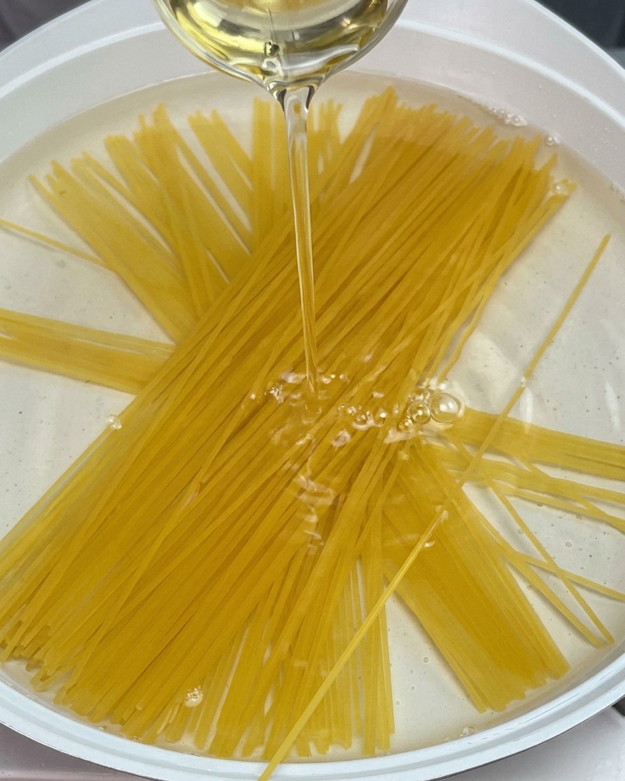 Trockene Spaghetti in einem Topf mit Wasser, über die ein Strahl Öl gegossen wird.