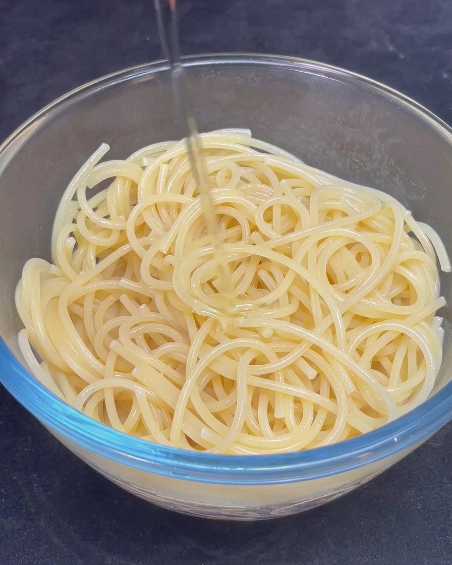 Ein Strahl goldenes Olivenöl wird über die gekochten Spaghetti in einer Glasschüssel gegossen.