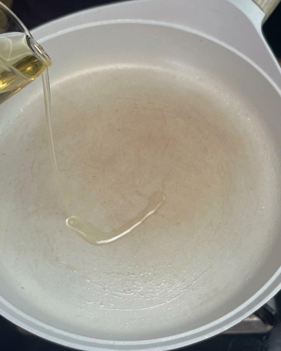 Speiseöl wird aus einem kleinen Krug in eine vorgeheizte weiße Pfanne gegossen.