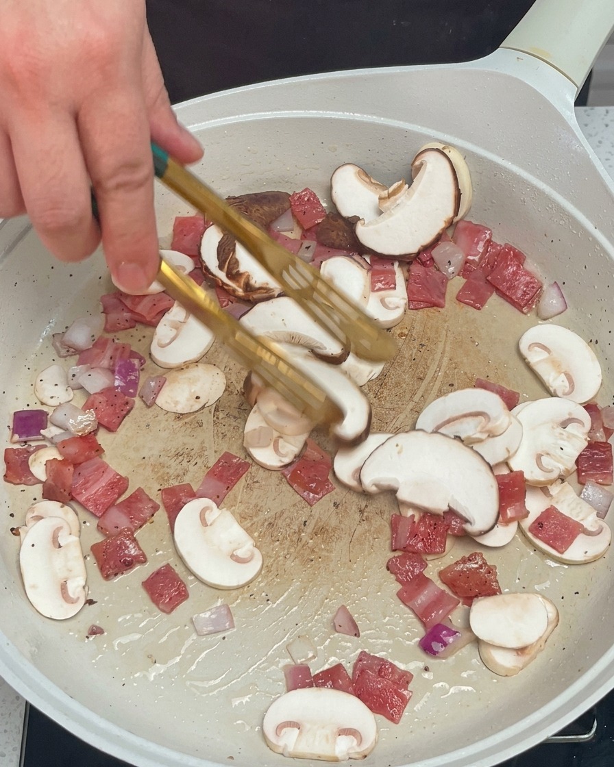 Pilze, Zwiebeln und Speck werden mit einer Grillzange in der Pfanne gewendet.