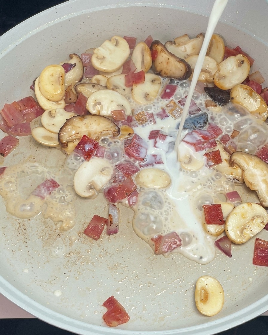 Weiße Milch wird zu den angebratenen Pilzen und dem Speck in die Pfanne gegossen.