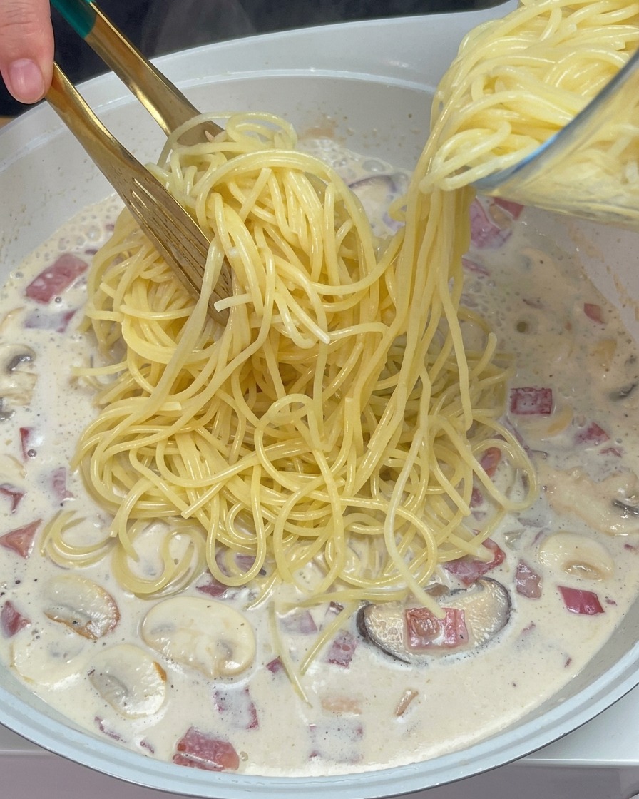 Gekochte Spaghetti werden in die köchelnde Milchsauce mit Pilzen und Speck gegeben.