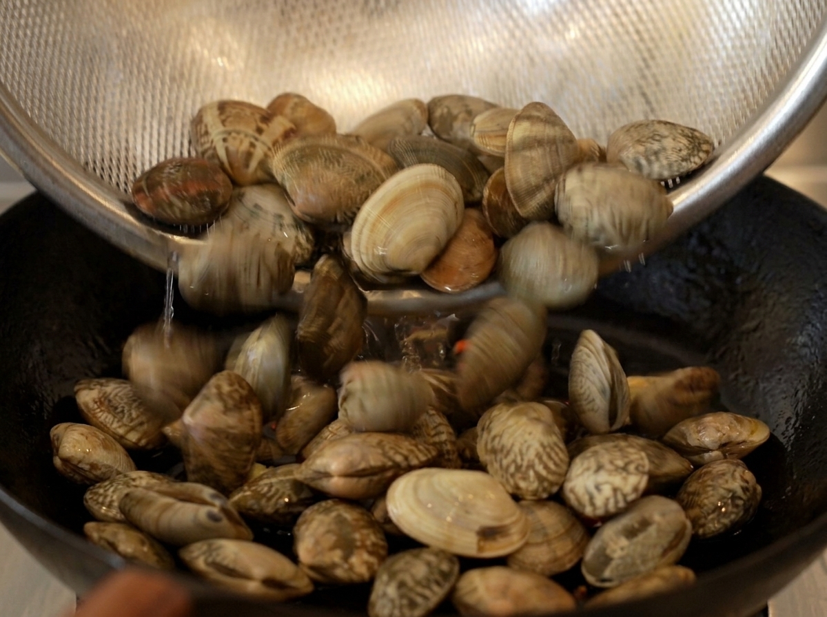 Gereinigte Venusmuscheln werden aus einem Sieb in eine heiße Pfanne geschüttet.