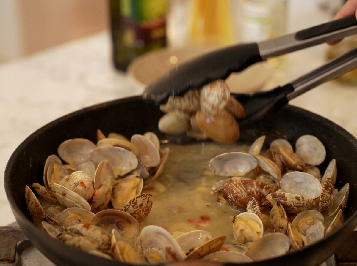 Küchenzange nimmt geöffnete Muscheln aus dem köchelnden Sud in der Pfanne.