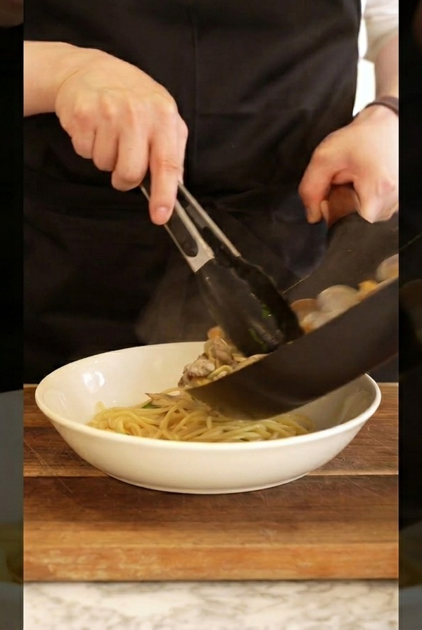 Ein Koch richtet die Muschel-Spaghetti mit einer Zange in einer weißen Schale an.