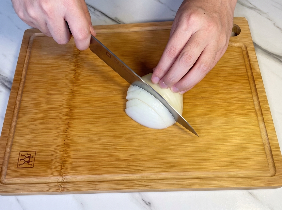 Hände würfeln mit einem Kochmesser eine halbe weiße Zwiebel auf einem Holzschneidebrett.