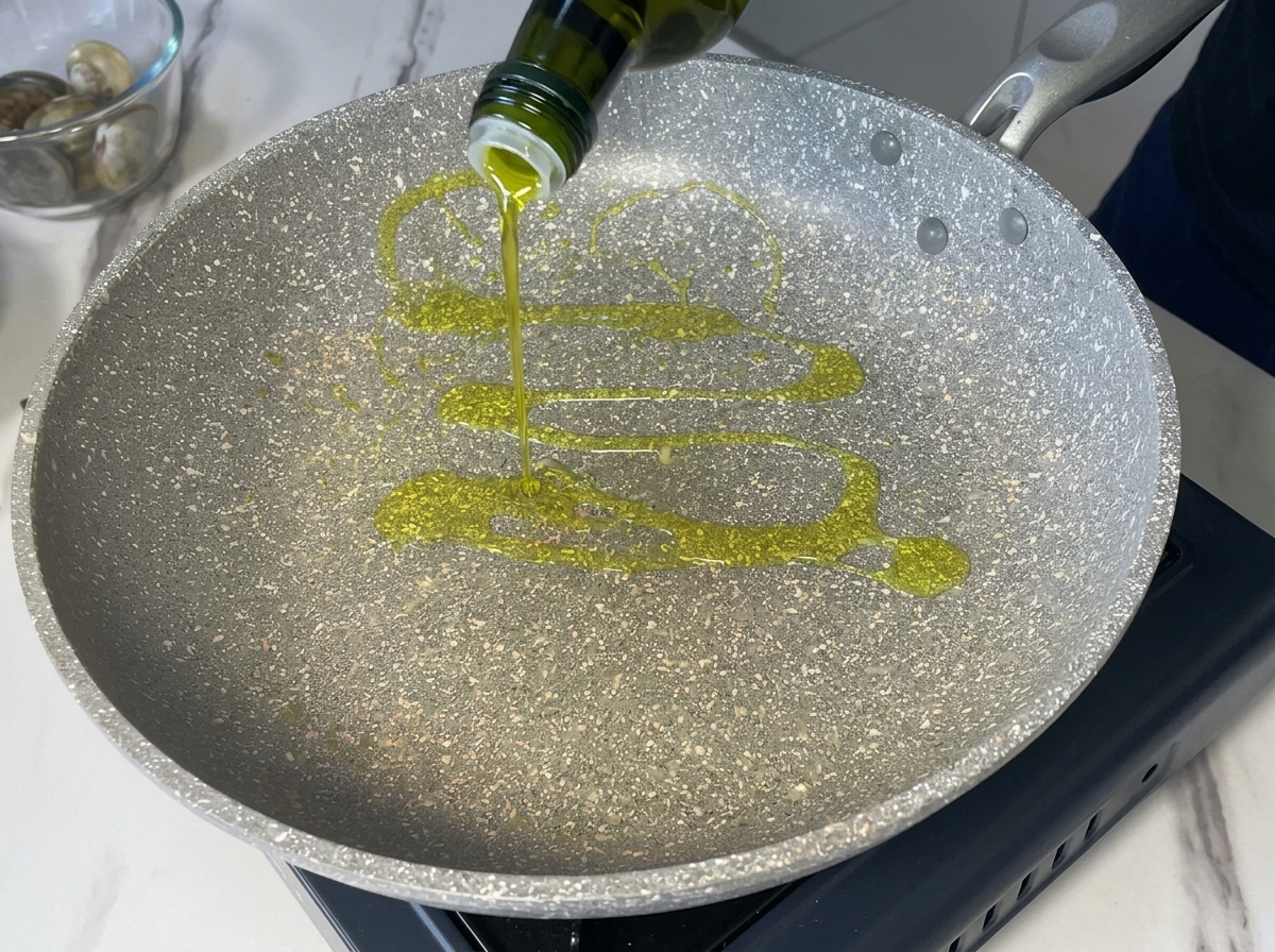 Natives Olivenöl extra wird aus einer dunkelgrünen Flasche in eine grau gesprenkelte Pfanne gegossen.