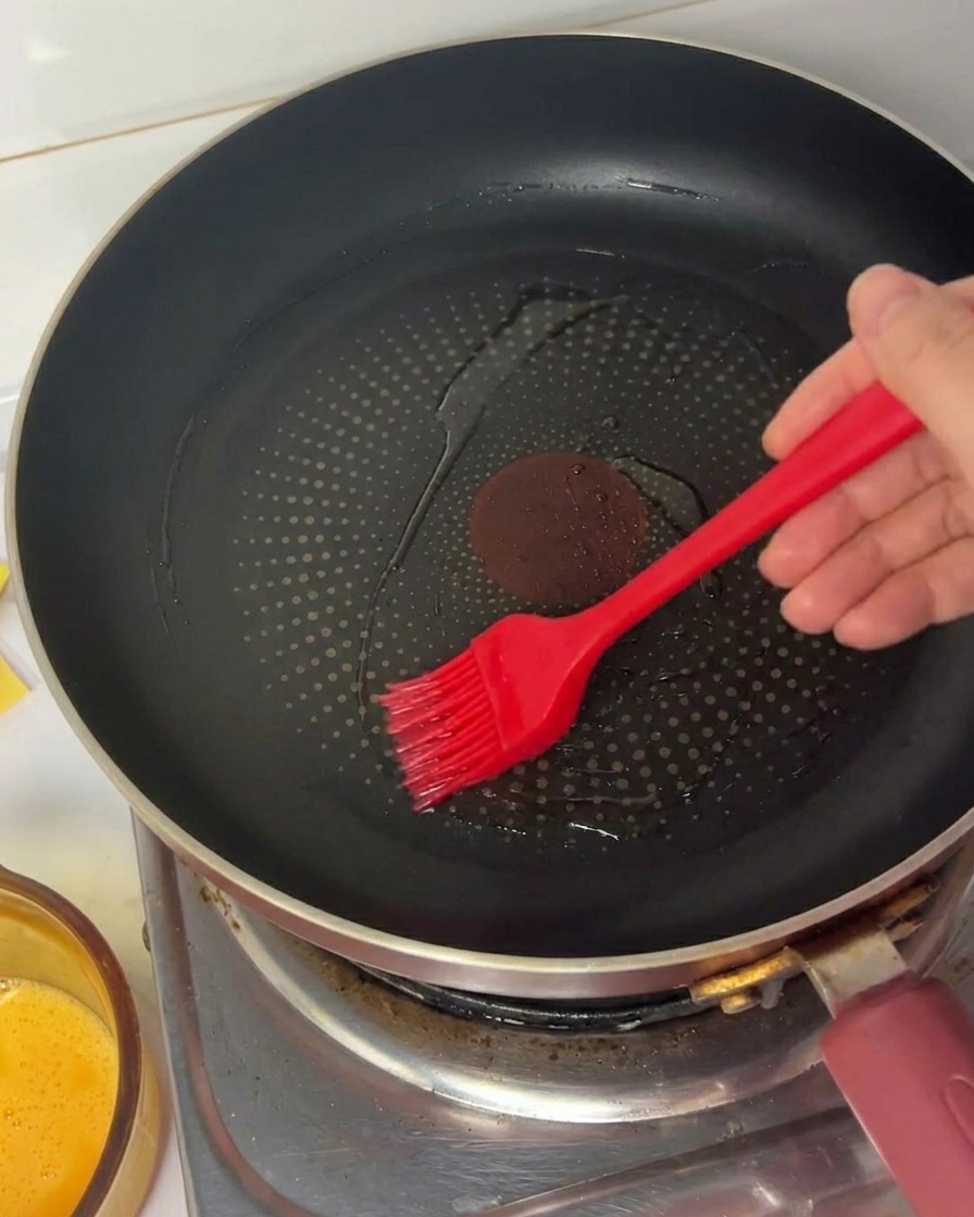 Eine Hand verteilt mit einem roten Silikonpinsel Speiseöl in einer schwarzen beschichteten Bratpfanne.