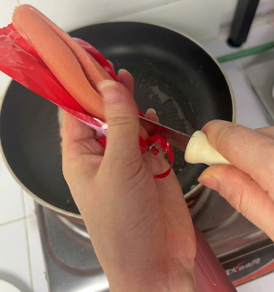Hände öffnen mit einem Messer die rote Verpackung einer Schinkenwurst über einer Pfanne mit bratendem Speck.
