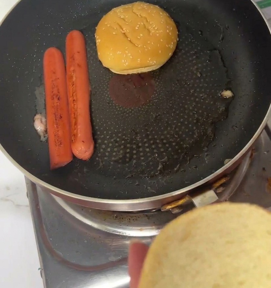 Sesam-Burgerbrötchen werden mit der Schnittfläche nach unten in einer Pfanne neben fertig gebratenen Würstchen geröstet.