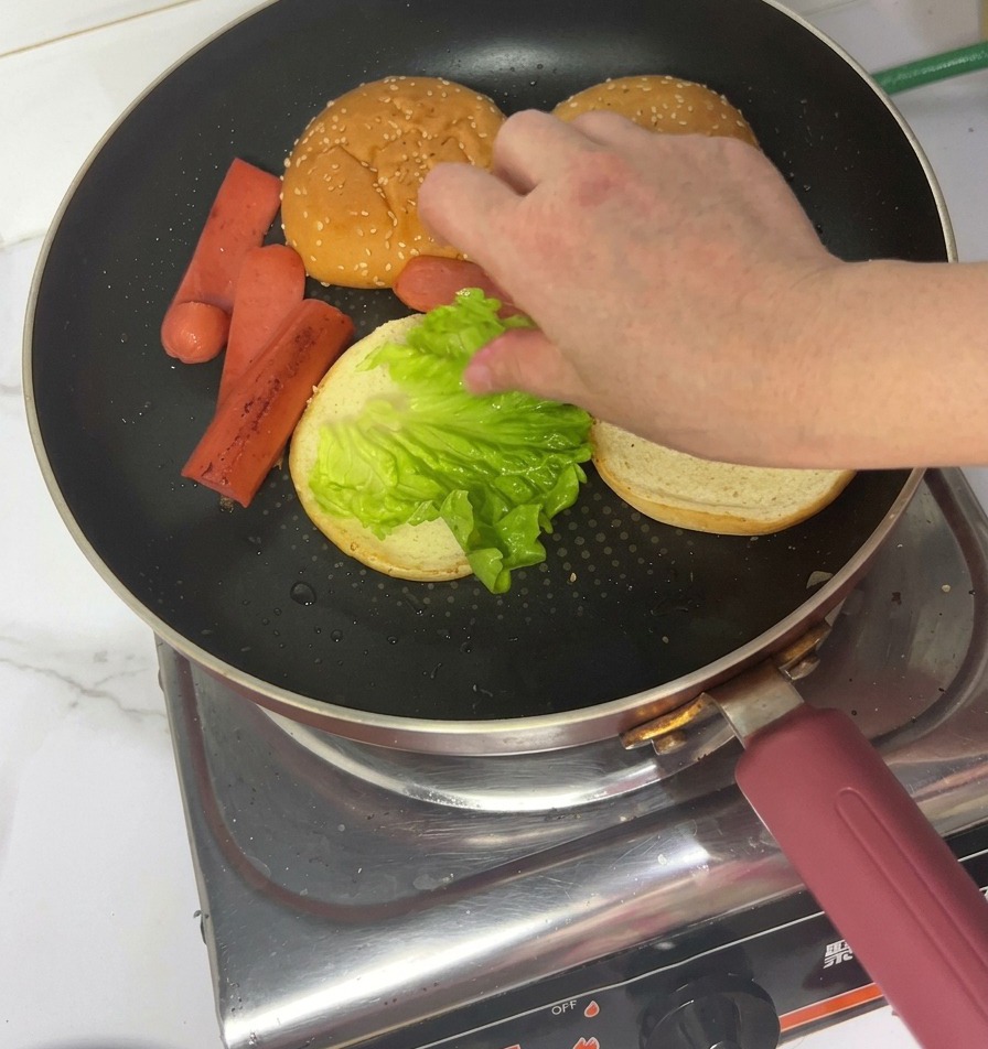 Eine Hand legt ein frisches, grünes Salatblatt auf eine geröstete Brötchenhälfte in einer Pfanne.