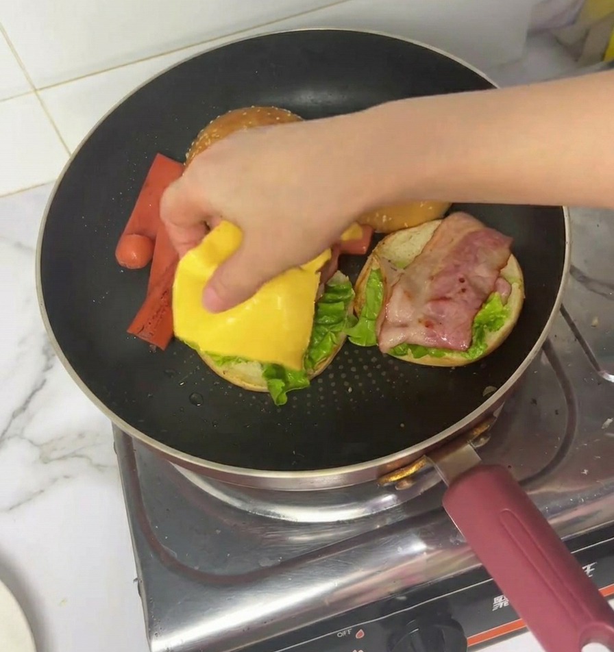 Eine Hand legt eine quadratische Scheibe gelben Käse auf einen halbfertigen Burger mit Salat und Speck in einer Pfanne.