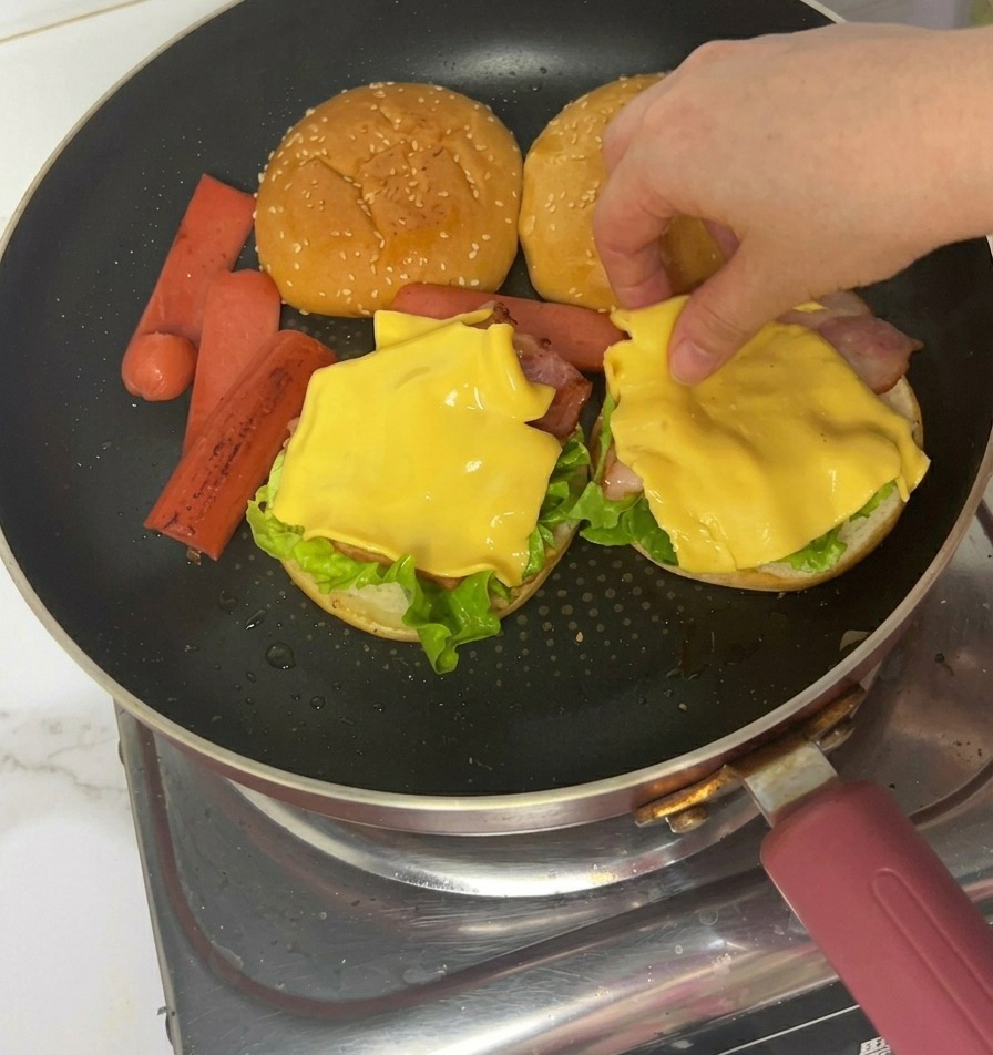 Hände legen mit einer Zange gebratene Schinkenwurstscheiben auf die Käseschicht eines teilweise belegten Burgers.