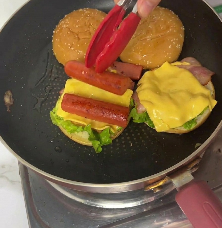 Ein Viertel eines dünnen Omeletts wird mit einer roten Zange in einer Bratpfanne auf den Burger gelegt.