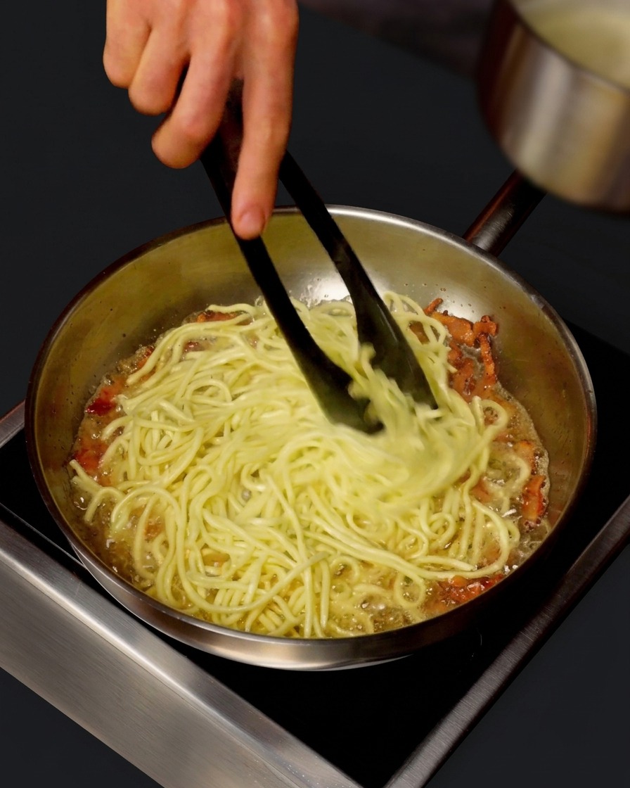 Ein kleiner Topf gießt heißes Nudelwasser in eine Pfanne mit Spaghetti, während diese mit einer Zange gewendet werden.