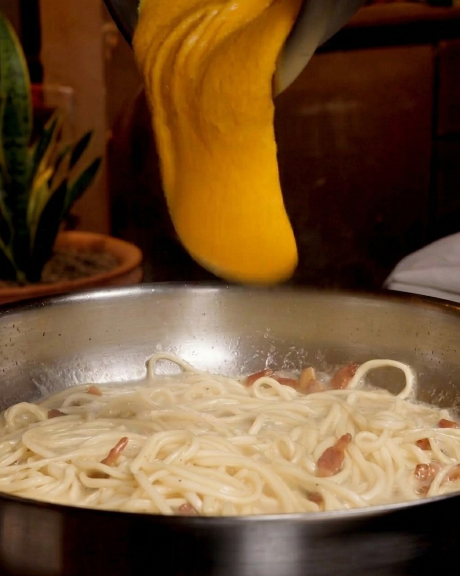 Eine dicke, leuchtend orangefarbene Ei-Käse-Mischung wird aus einer Metallschüssel über die Spaghetti in der Pfanne gegossen.