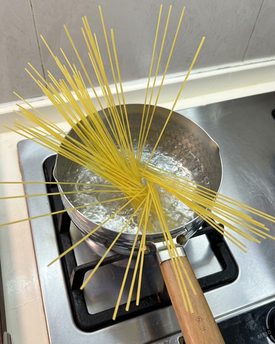 Trockene Spaghetti, die fächerförmig in einem kleinen Topf mit kochendem Wasser auf einem Gasherd stehen.