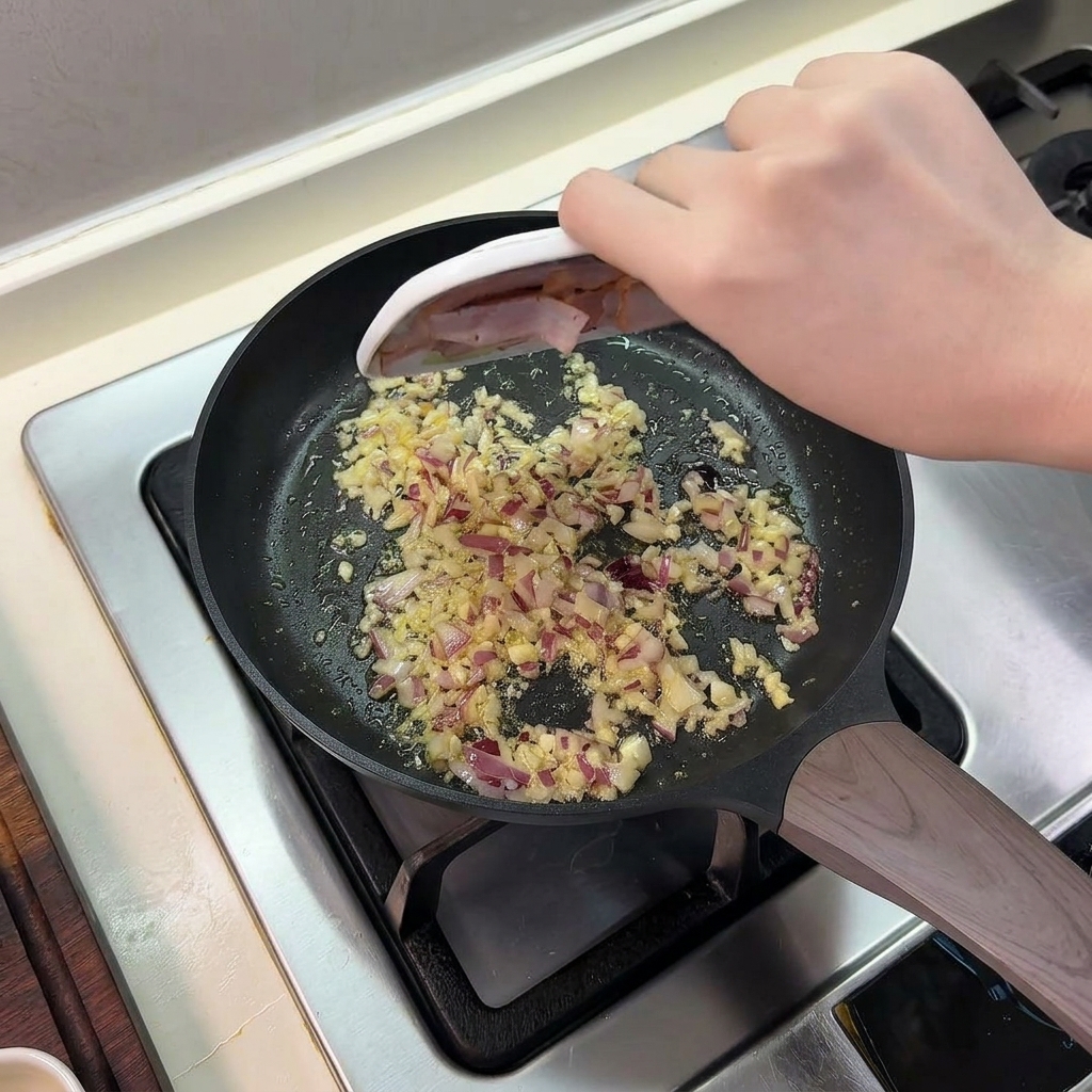 Geschnittener Speck, der von einem kleinen Teller in eine Bratpfanne mit angeschwitzten Zwiebeln und Knoblauch rutscht.