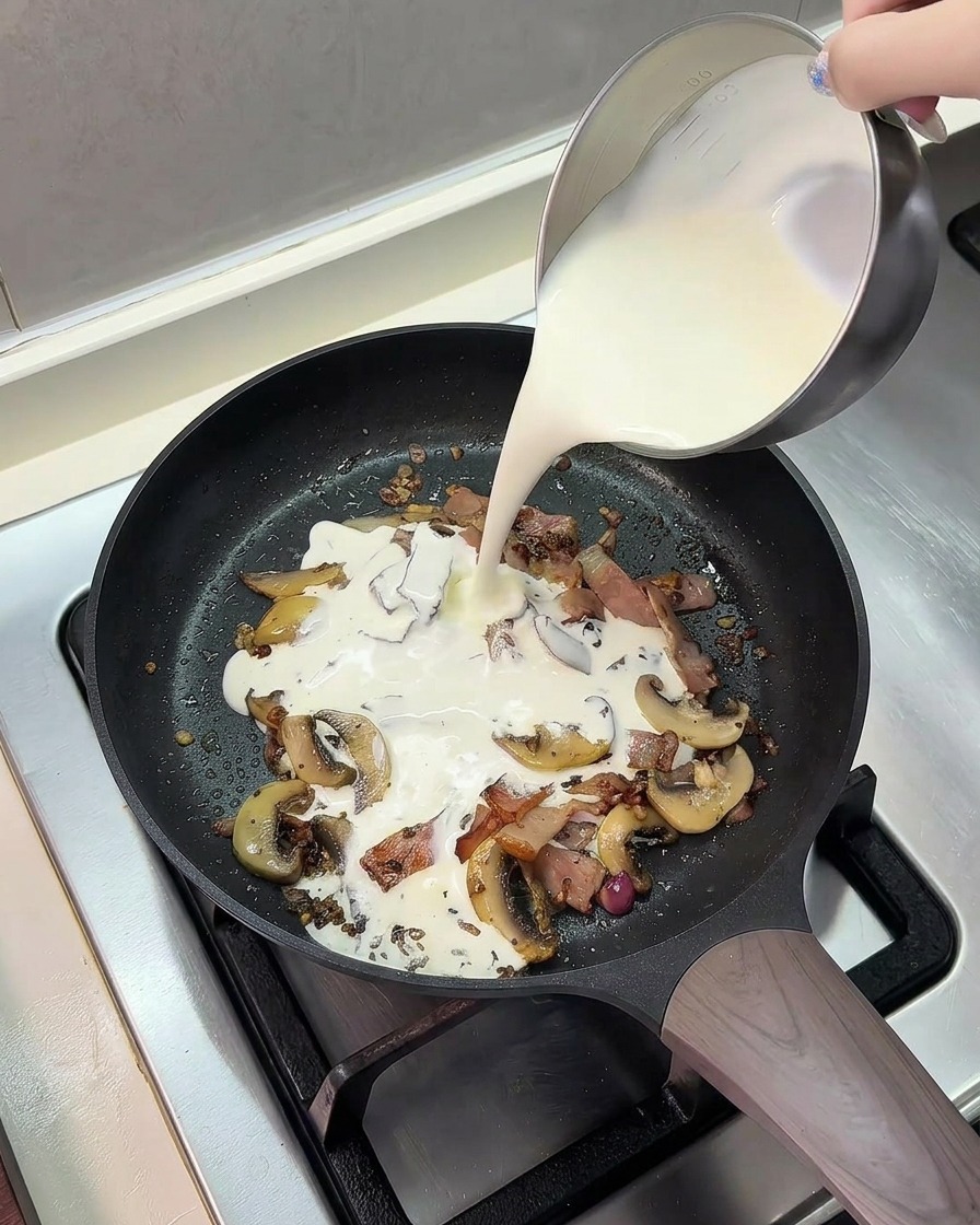 Eine Mischung aus Milch und Sahne wird aus einem Metallkännchen in eine Bratpfanne mit gebratenem Speck und Pilzen gegossen.