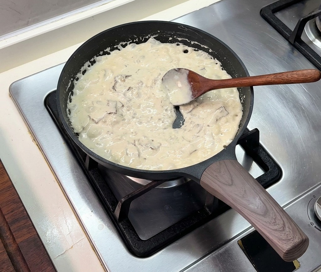 Eine dicke, blubbernde Sahnesauce, die in einer schwarzen Bratpfanne köchelt und mit einem Holzlöffel umgerührt wird.