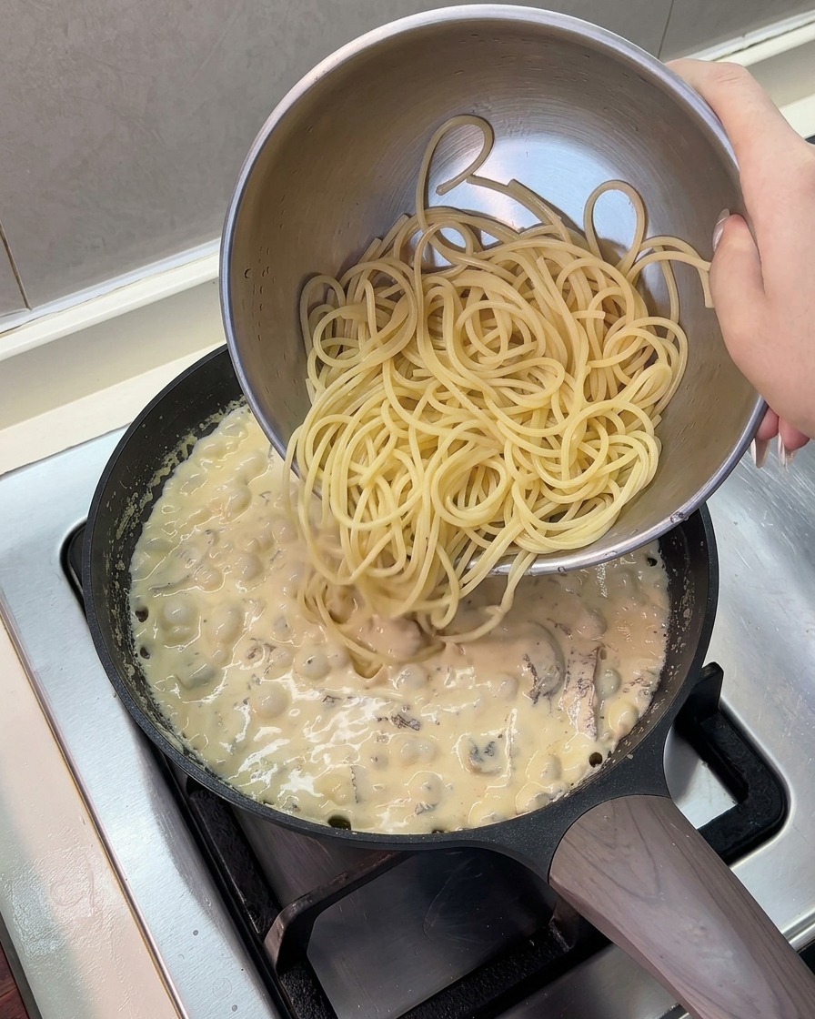 Gekochte Spaghetti-Nudeln werden aus einer Edelstahlschüssel in eine Pfanne mit köchelnder Sahnesauce geschüttet.