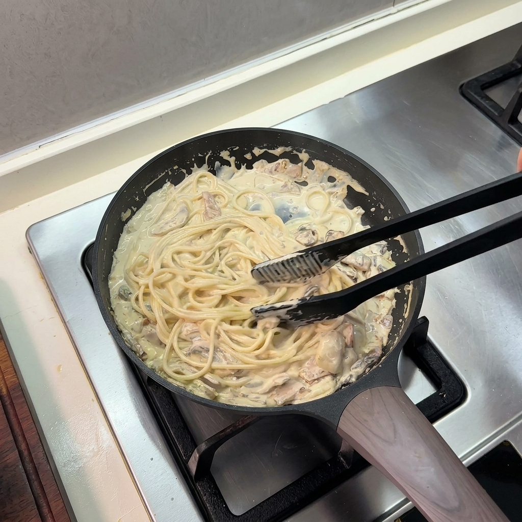 Eine schwarze Küchenzange wendet gekochte Spaghetti in einer reichhaltigen Sahnesauce in einer Bratpfanne.