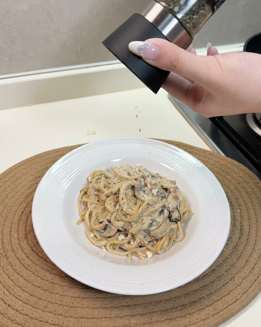 Eine Hand reibt frischen Käse über einen warmen Teller mit cremigen Pilz-Speck-Spaghetti und streut Kräuter darüber.