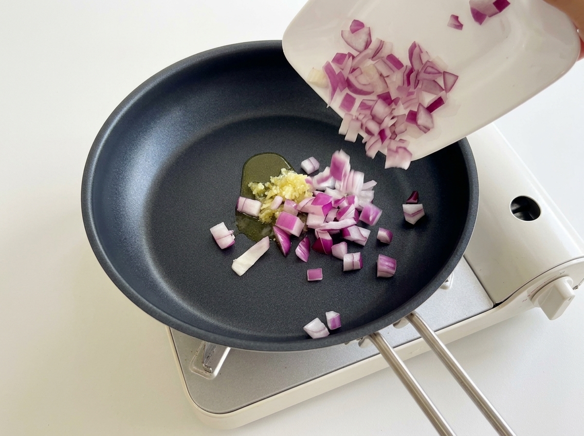 Gewürfelte rote Zwiebeln werden in eine Pfanne mit bereits angeschwitztem Knoblauch gegeben.