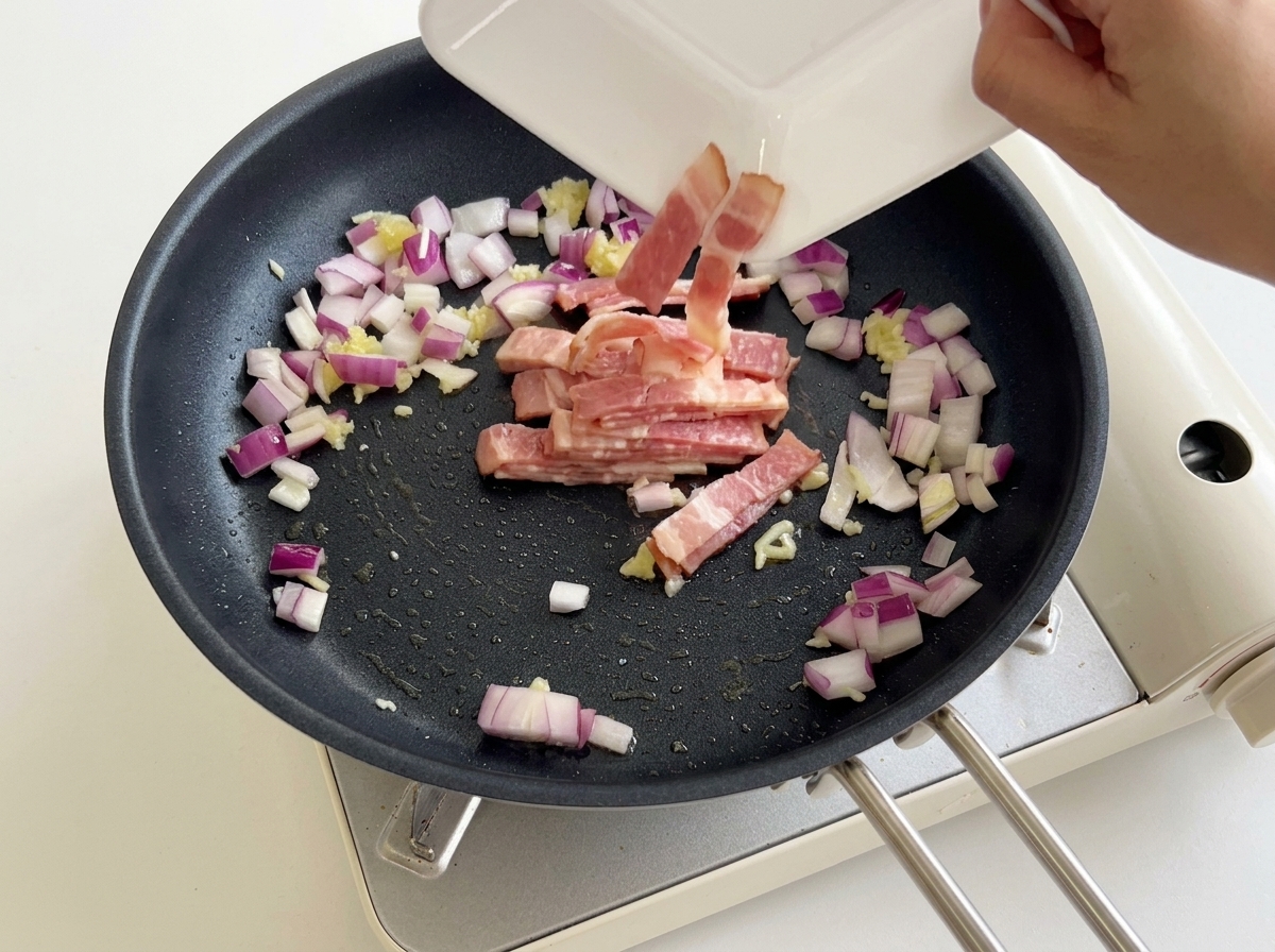 Geschnittener Speck wird von einem weißen Teller in die Pfanne zu den Zwiebeln und dem Knoblauch gegeben.