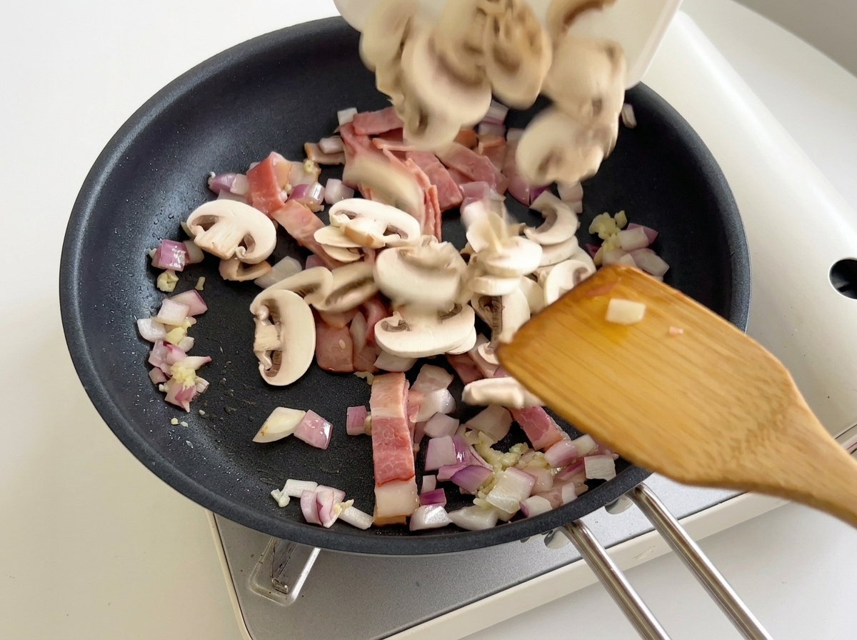 Champignonscheiben werden in die Pfanne mit Speck und Zwiebeln gegeben.