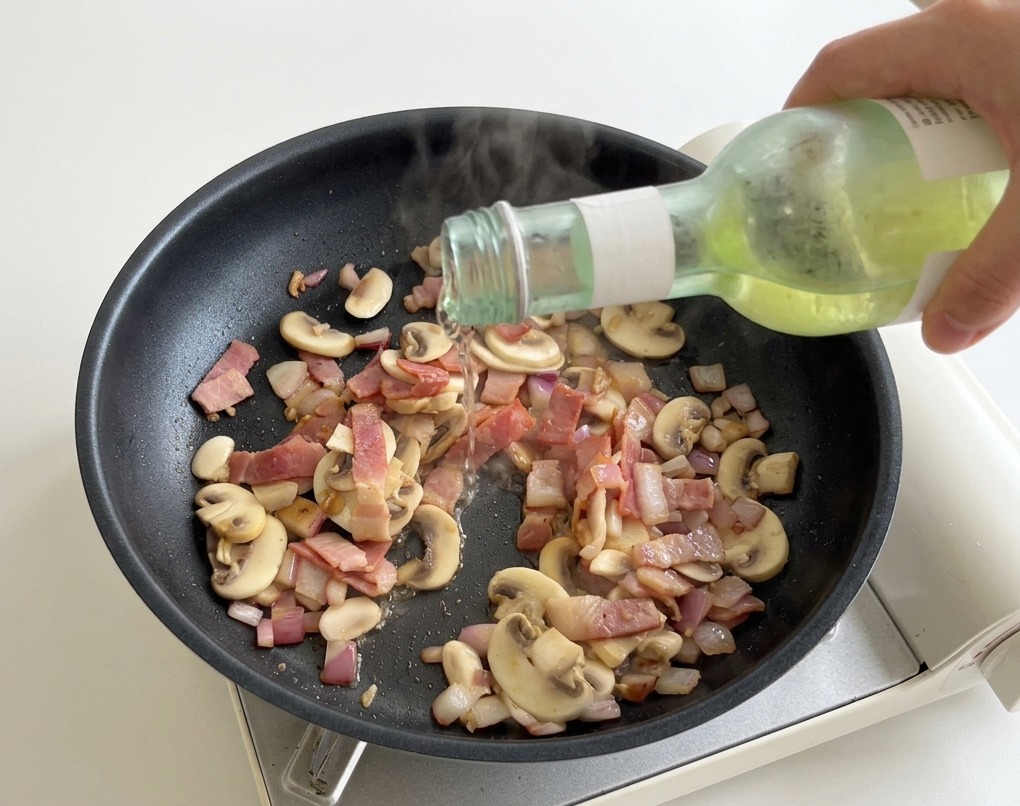 Ein Schuss Weißwein wird aus einer kleinen grünen Flasche in die heiße Pfanne mit Pilzen und Speck gegossen.
