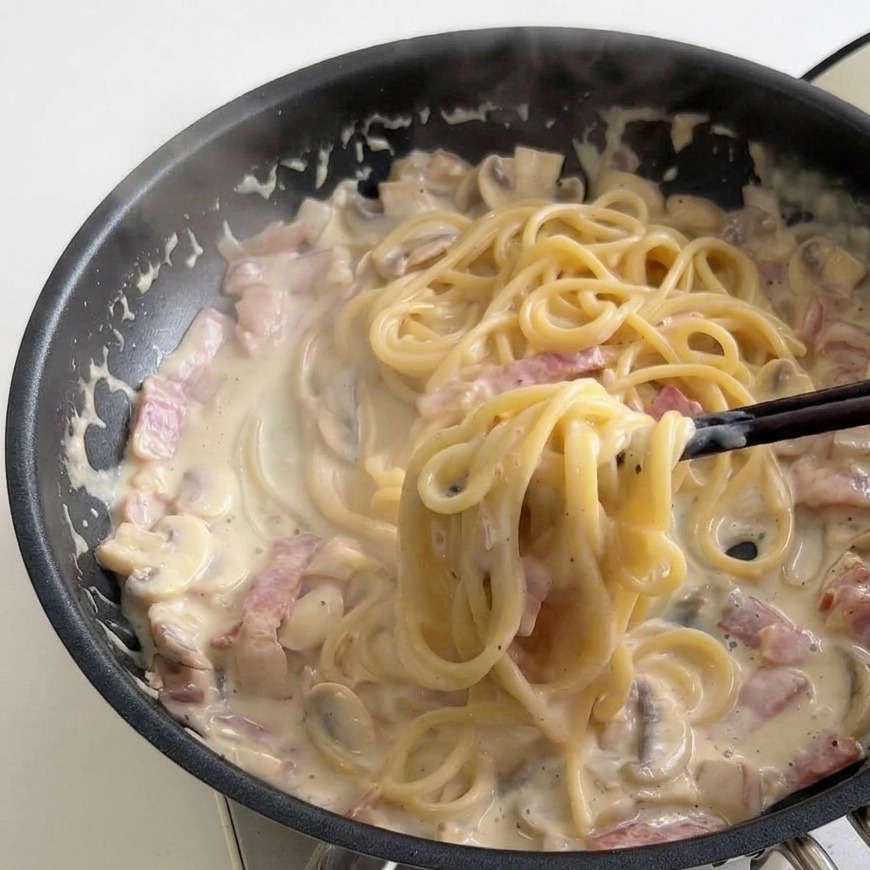Stäbchen drehen Spaghetti in einer Pfanne mit sämiger, blubbernder Milch-Sauce.