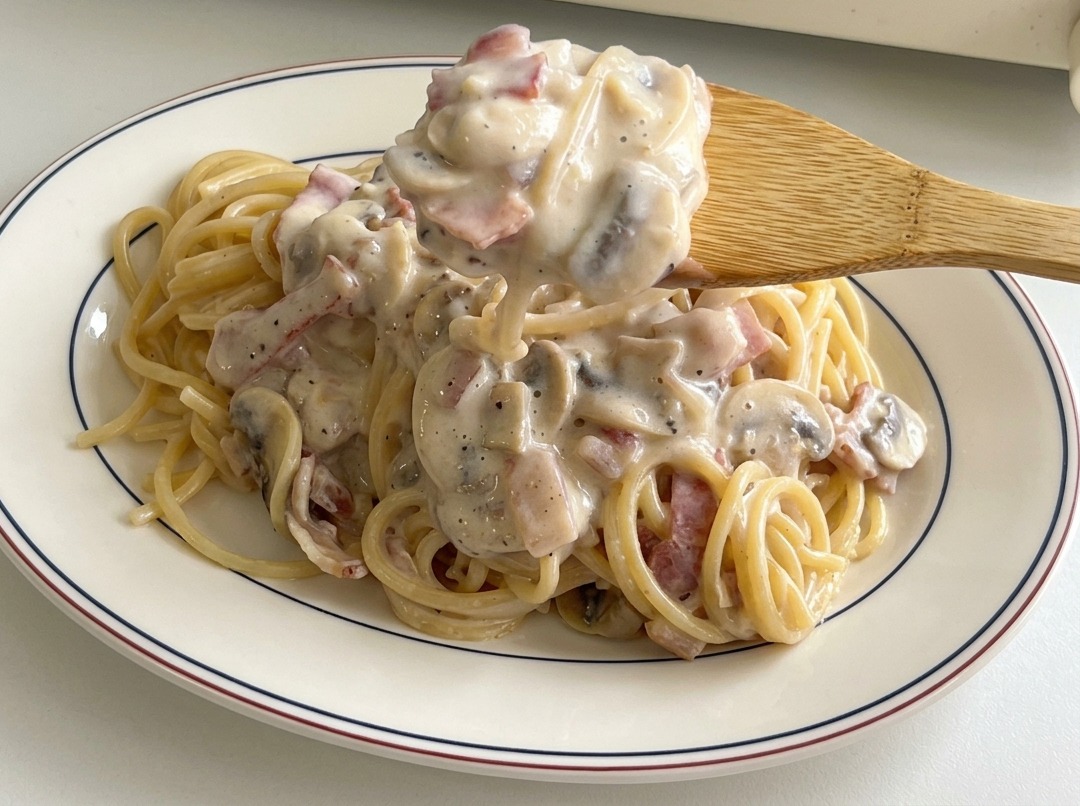 Ein Holzlöffel gibt die restliche Sauce mit Speck und Pilzen über die angerichteten Spaghetti.