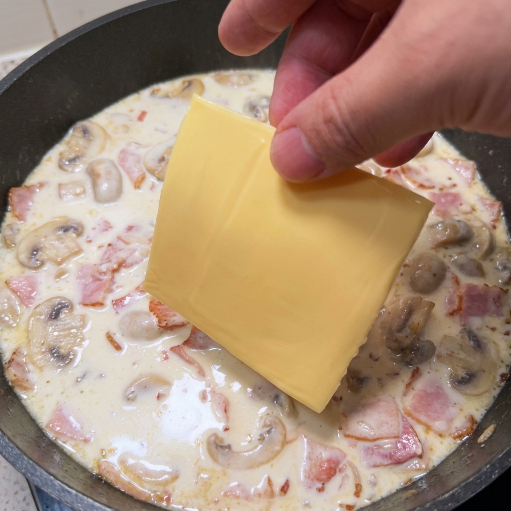 Eine Hand legt eine Scheibe gelben Käse in die Pfanne mit der cremigen Sauce.