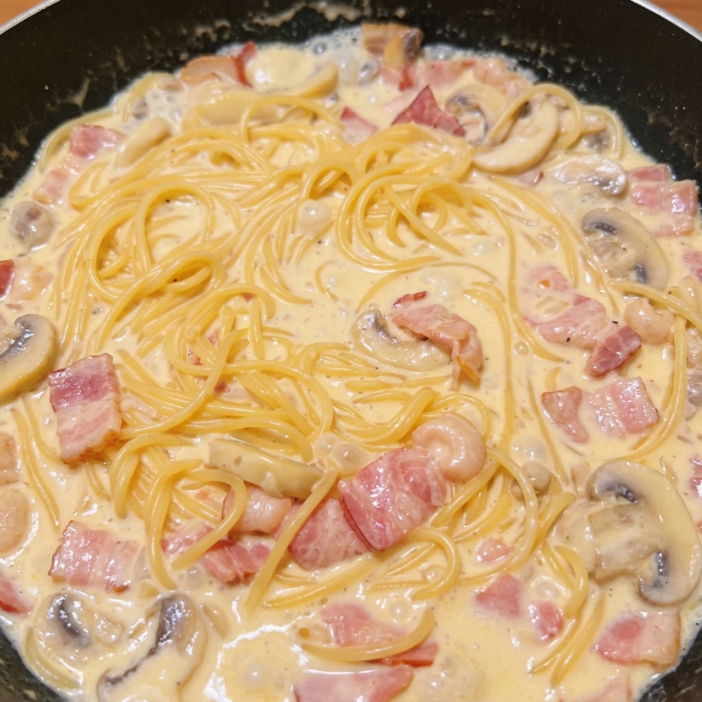 Gekochte Spaghetti werden in der Pfanne mit der Speck-Pilz-Rahmsauce vermengt.