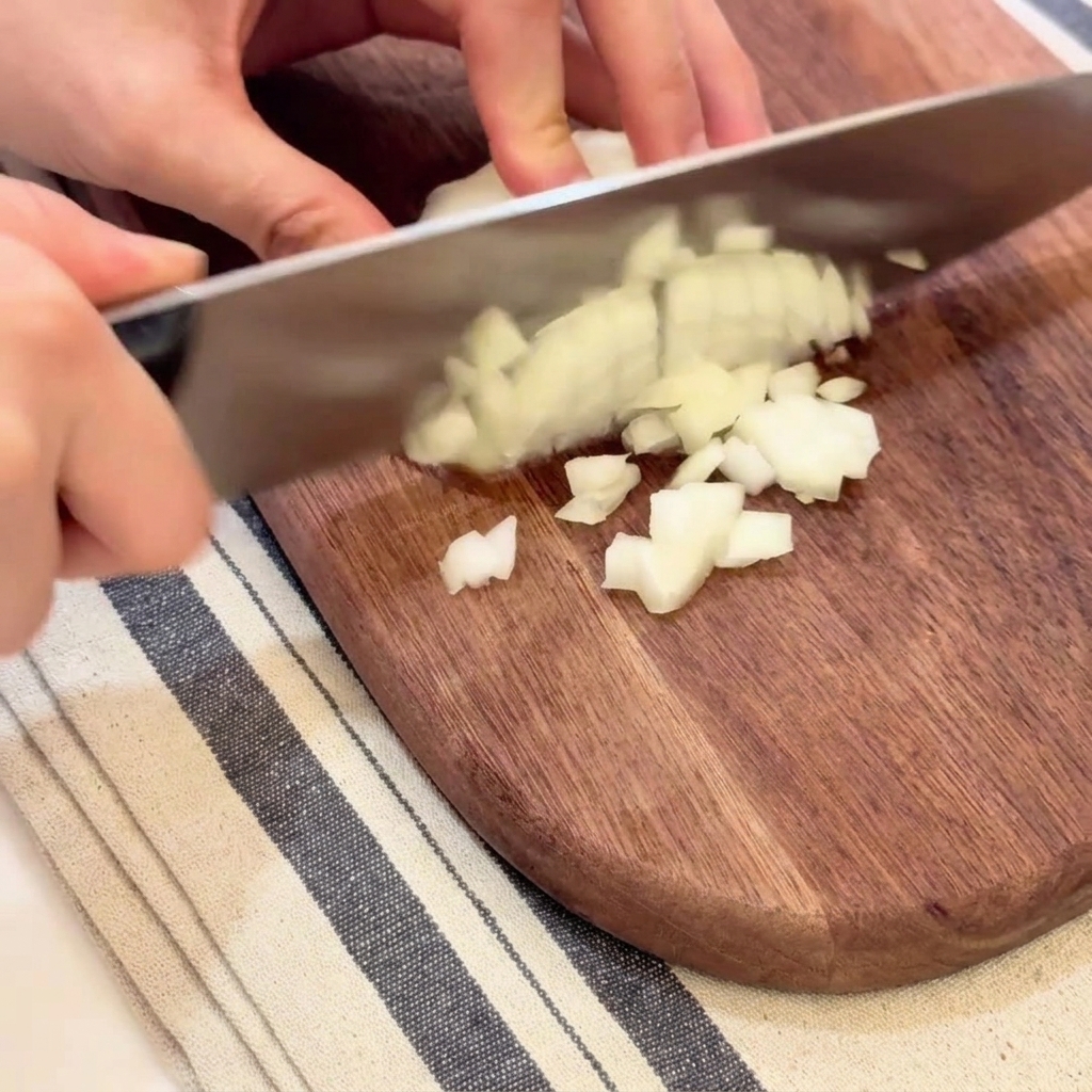 Hände würfeln eine weiße Zwiebel vorsichtig in kleine, gleichmäßige Stücke auf einem Schneidebrett.