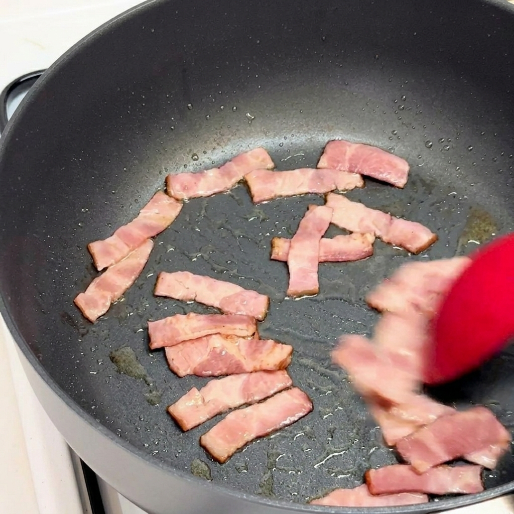 Speckstücke werden mit einem roten Pfannenwender in einer schwarzen Pfanne angebraten.