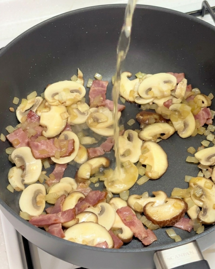 Eine klare Flüssigkeit wird in die Pfanne mit Pilzen, Speck und Zwiebeln gegossen.