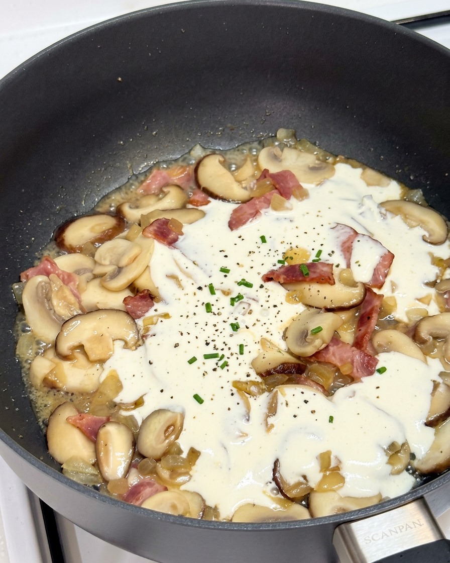 Sahne köchelt in einer Pfanne mit Pilzen und Speck, gewürzt mit Pfeffer und Kräutern.