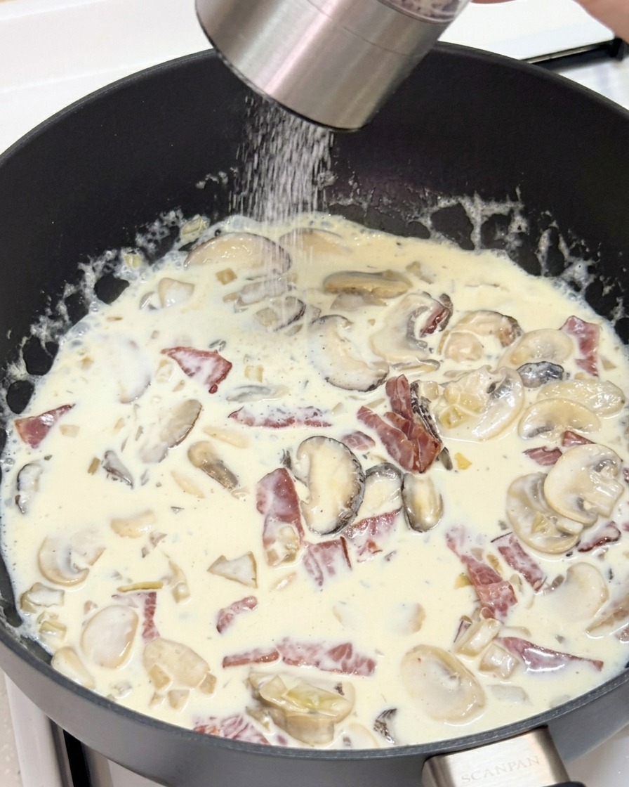 Frischer schwarzer Pfeffer wird über die cremige Pilz-Speck-Sauce gemahlen.