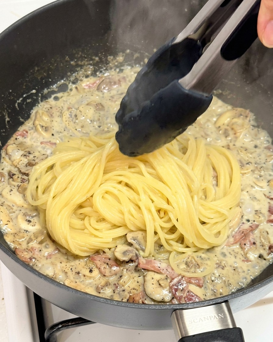 Eine Küchenzange gibt frisch gekochte Spaghetti in die Pfanne mit der Trüffel-Pilz-Sauce.